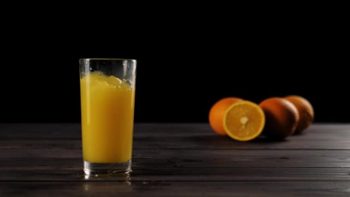 冰块落在高高的玻璃杯中，上面放着新鲜的橙汁或菠萝汁，在木制桌子上产生飞溅和气泡，上面装饰着橙子，在黑