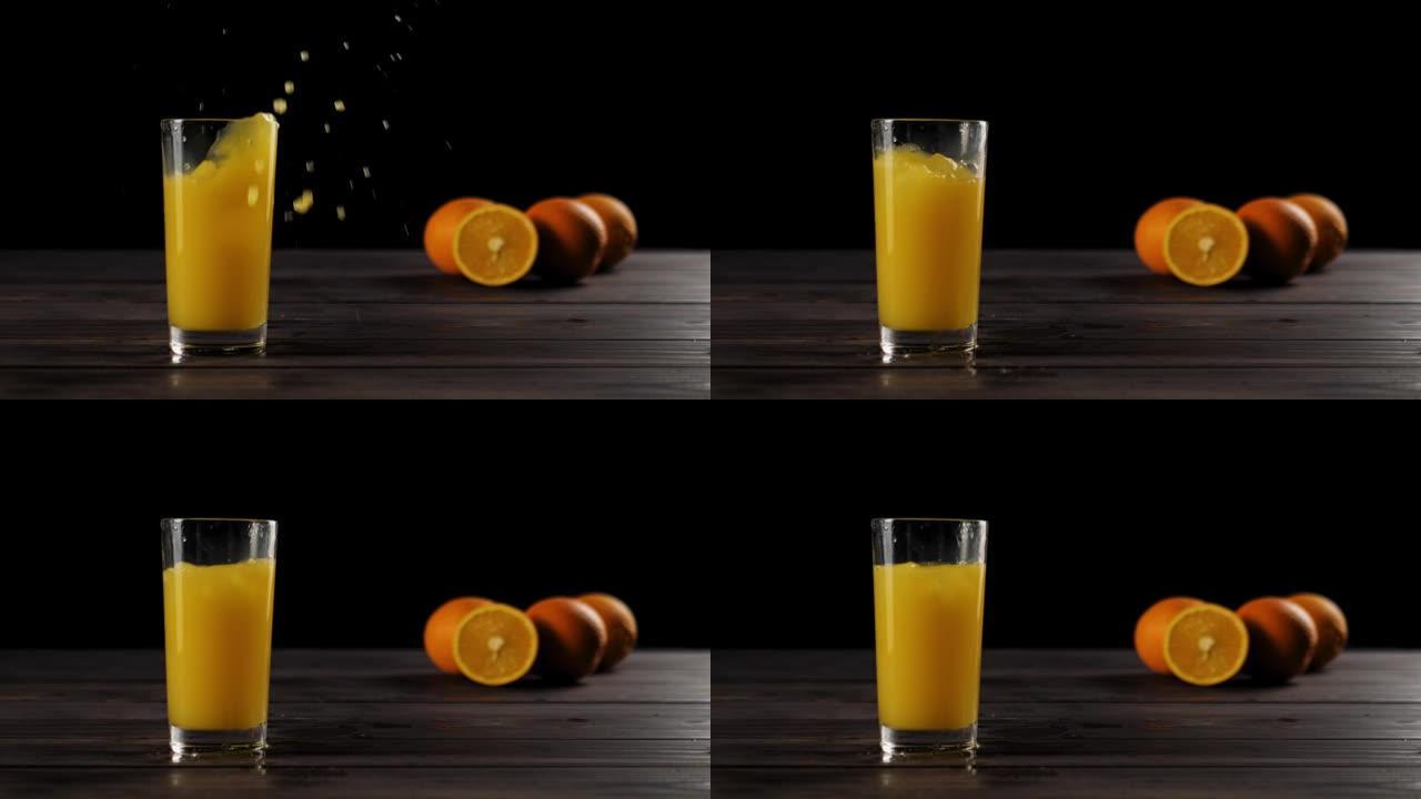 冰块落在高高的玻璃杯中，上面放着新鲜的橙汁或菠萝汁，在木制桌子上产生飞溅和气泡，上面装饰着橙子，在黑