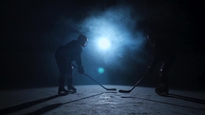 两名男子曲棍球运动员穿着制服，戴着头盔，手里拿着棍子，在冰上滑冰的深色轮廓。一名运动员在黑暗竞技场的