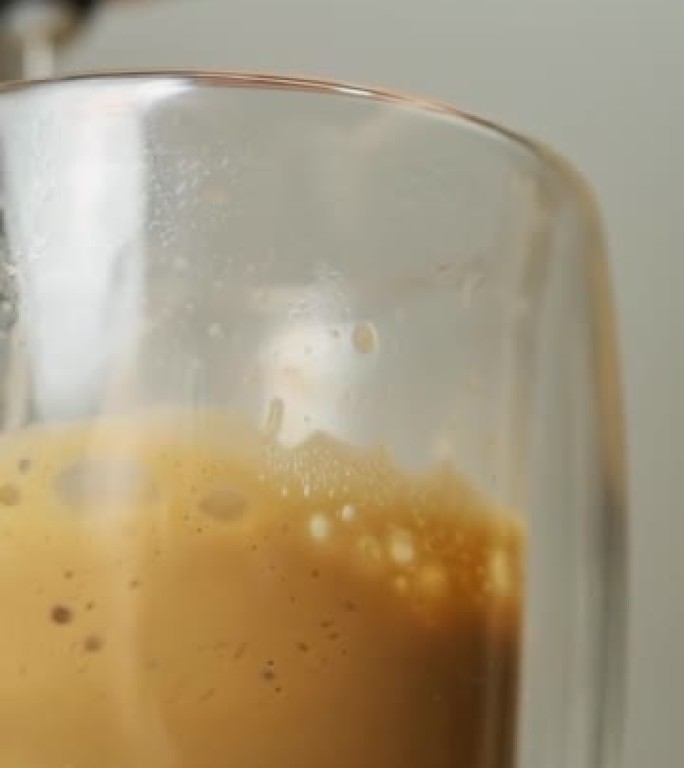 在双层玻璃卡布奇诺玻璃中，用电动起泡剂摇动牛奶泡沫。在家准备咖啡饮料。垂直视频