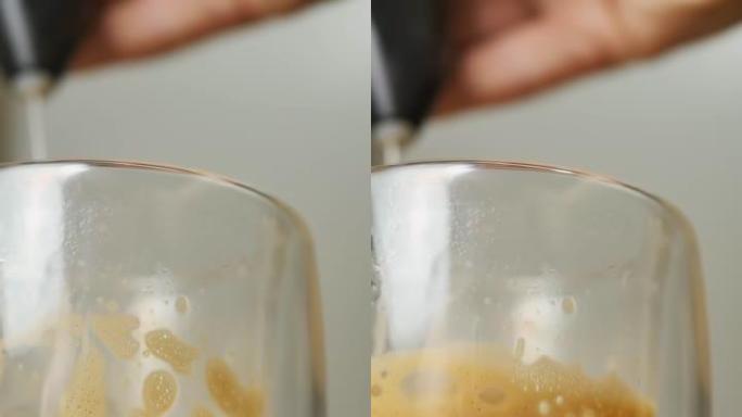 在双层玻璃卡布奇诺玻璃中，用电动起泡剂摇动牛奶泡沫。在家准备咖啡饮料。垂直视频
