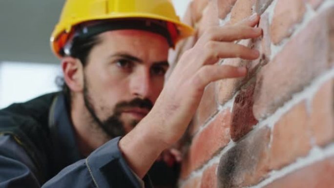 在镜头前的特写镜头中，富有魅力和好看的建筑工人从新墙中分析了红砖的结构，他戴着安全帽和制服。4k