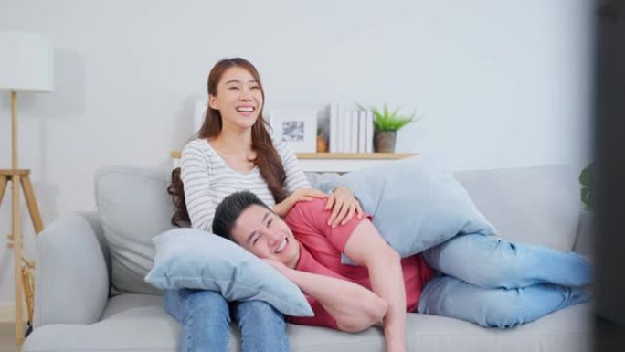 亚洲年轻的情侣在家里的客厅一起看电影。迷人的浪漫新婚男女坐在沙发上，看着电视节目，在家里一起欢笑。