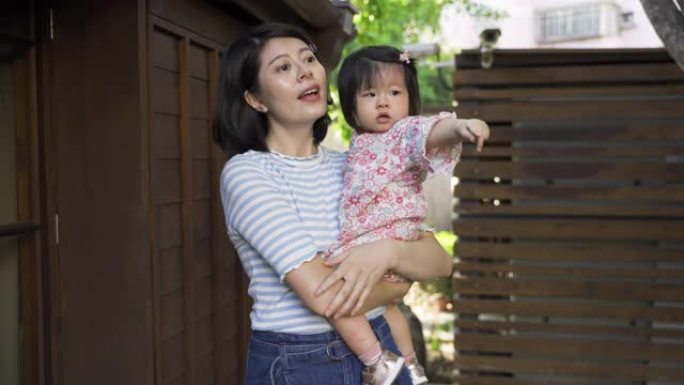 肖像拍摄开朗的日本母亲正在向怀抱中的蹒跚学步的女儿解释，好奇地指着花园中的远方。