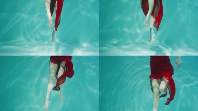 穿着红色雪纺服装跳舞的女人的水下镜头。女舞者在眩光的蓝色水柱中优雅地移动双腿。女子水下射击，慢动作特