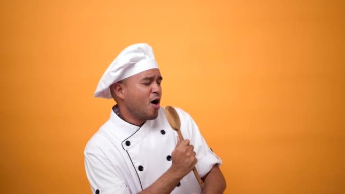 肖像快乐有趣的男人厨师拿着厨房用具跳舞和唱歌的音乐孤立在黄色背景上。