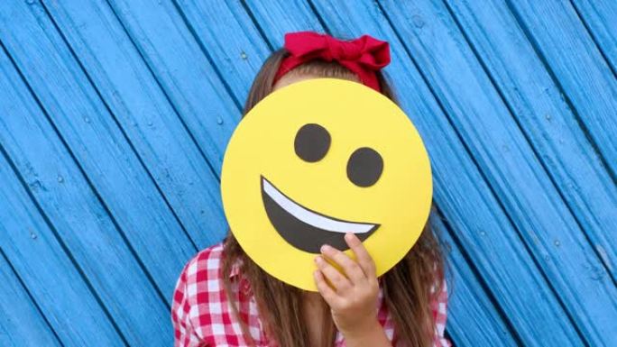女孩躲在幸福微笑表情的纸板脸后面。世界表情符号日。做自己的幸福