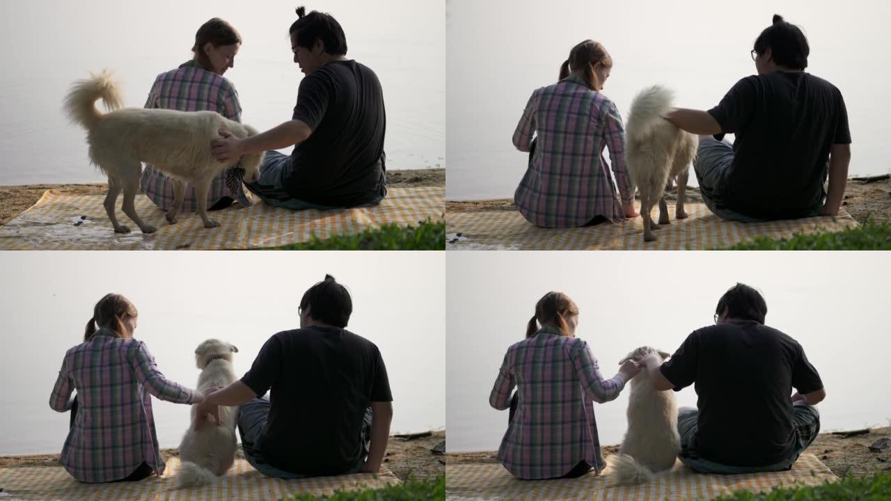 年轻夫妇坐在湖岸与狗闲逛。