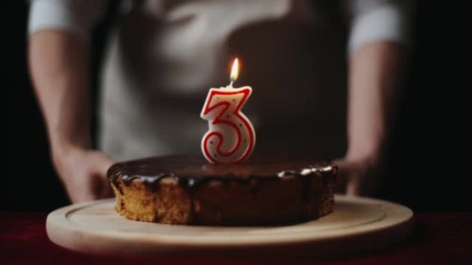 穿着厨房围裙的年轻女子将自制的巧克力蛋糕或带有3号蜡烛的馅饼放在家里的桌子上。三岁，庆祝生日概念