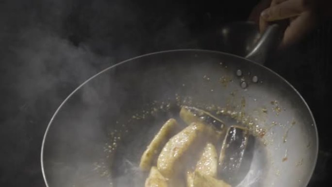 一家亚洲餐厅的厨师在热煎锅里炸茄子，把它们扔到厨房的背景下。男人用热香料，大蒜和糖浆在油中炸蔬菜。慢