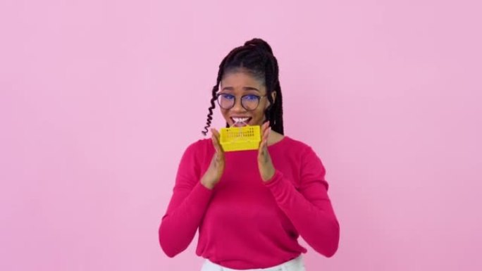 穿着粉色衣服的非洲裔美国女孩拿着一个迷你玩具购物篮。少女家庭主妇初学者站在坚实的粉红色背景上