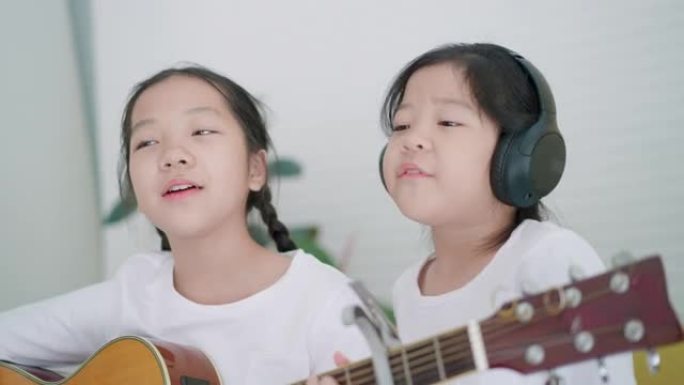 快乐时光在音乐教室里，7岁的小学老年亚洲女孩表演弹吉他，和5岁的小学老年亚洲女孩一起坐着，戴着无线耳