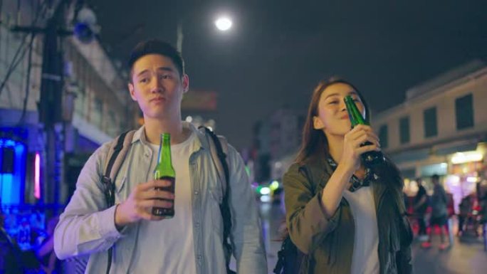 亚洲迷人的夫妇喝酒，一起开派对。在城市旅行的年轻男女游客在旅途中度过假期，在漆黑的夜晚一起拿着一瓶啤