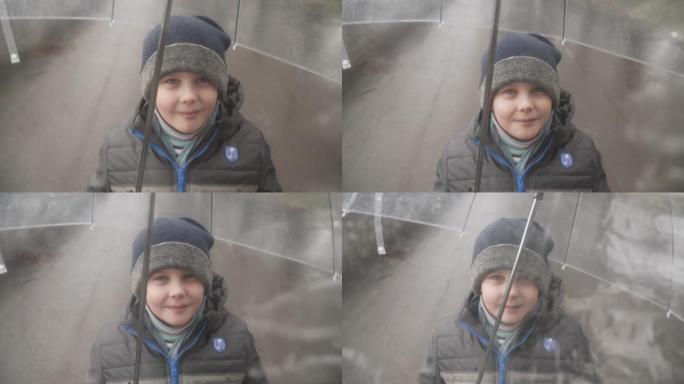 穿着夹克和帽子的可爱男孩透过潮湿透明的雨伞看和微笑。