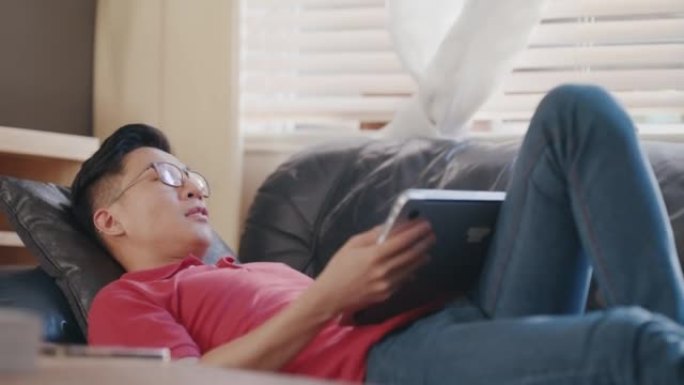 亚洲男子躺在家里的沙发上，肚子上放着一台笔记本电脑。年轻人下班后在房子里放松。中国男子在公寓里很舒服