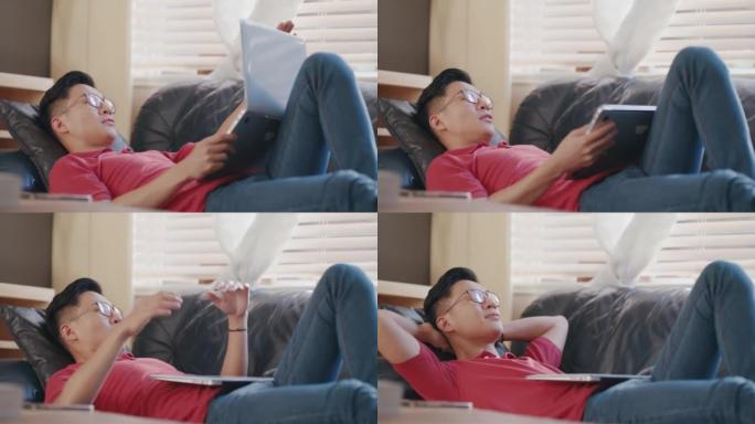 亚洲男子躺在家里的沙发上，肚子上放着一台笔记本电脑。年轻人下班后在房子里放松。中国男子在公寓里很舒服