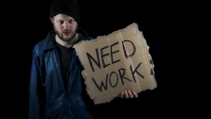 无家可归者拿着一张写着“需要工作”的纸板。穿着脏衣服的男流浪汉，黑色背景。难民正在找工作。贫困。大胡