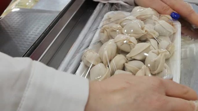 一名女杂货店工作人员的手将冷冻饺子包装成泡沫托盘，并用工业热包装机用食品拉伸膜包裹。手工制作。食品包