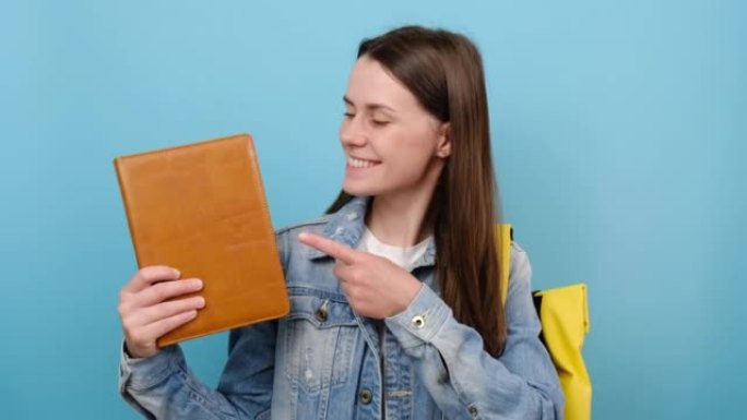 肖像快乐女孩青少年学生用手指指着书，露出拇指，穿着黄色背包，在工作室里孤立在蓝色背景上。高中教育大学