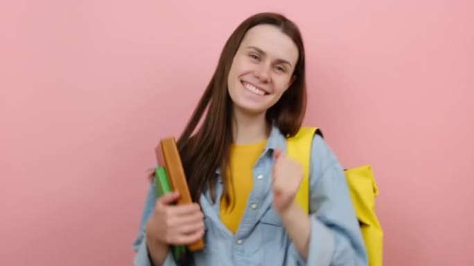 美丽的微笑女孩青少年学生拿着书跳舞休息，穿着衬衫和黄色背包，在粉色背景工作室中孤立地摆姿势。高中教育