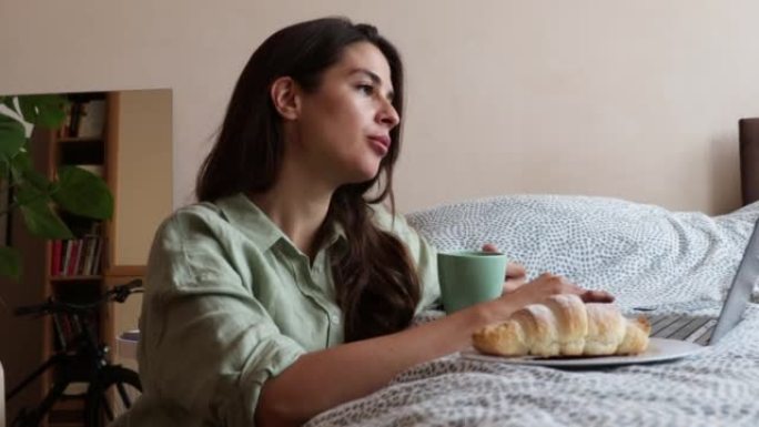 年轻的自由职业者的女商人喝第一杯咖啡有羊角面包作为早餐，并在她的笔记本电脑上工作。外籍女性早上在家里