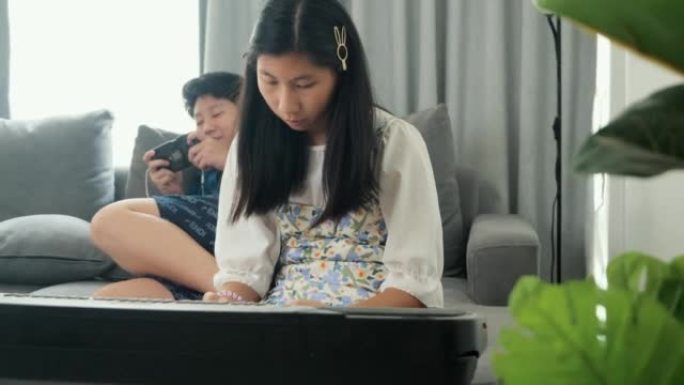 亚洲女孩在家里玩手机游戏的哥哥附近的沙发上通过手机应用学习玩键盘乐器，生活方式概念。