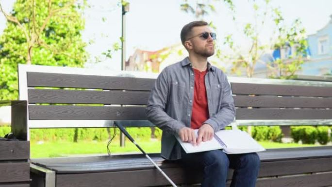 年轻的盲人坐在城市公园的长凳上阅读盲文书籍