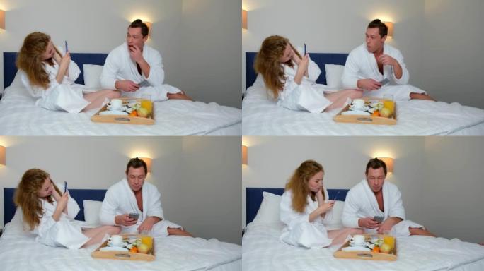 穿着浴袍的男人和女人在智能手机上拍照，在酒店的床上吃饭。