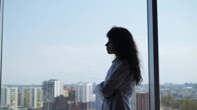 迷人的悲伤女医生的肖像，卷发透过窗户城市背景，感到疲倦。一位实习女医生的体贴剪影，对着窗户思考。