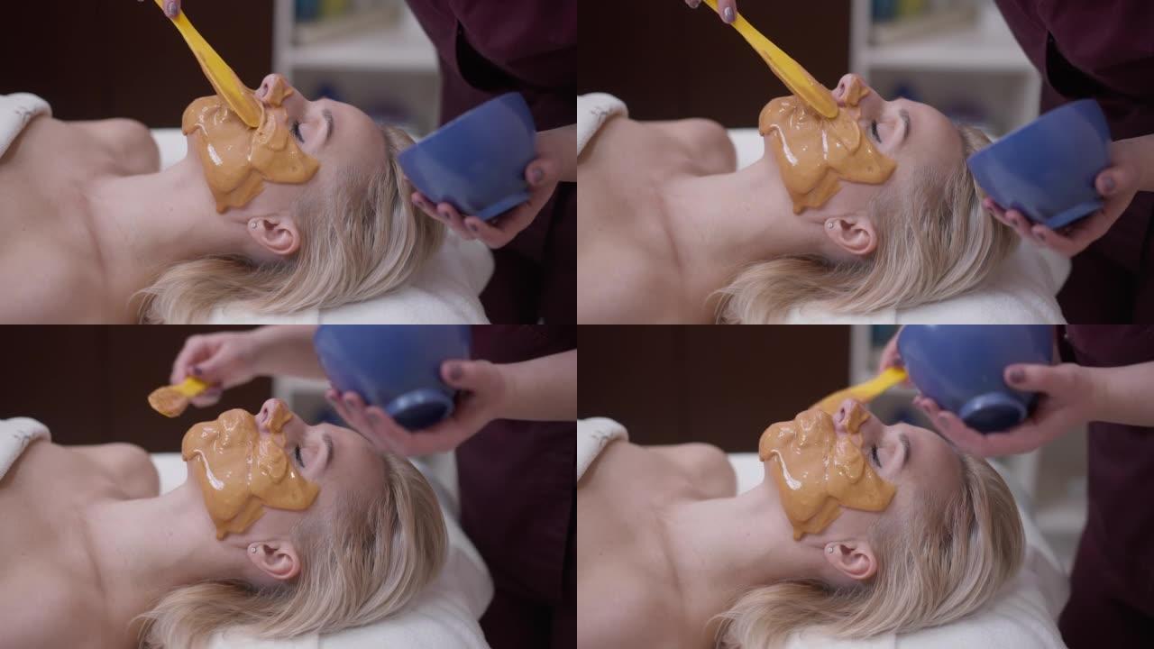 无法识别的美容师在美容水疗中心的客户脸上涂上黄色蜂蜜面膜。放松的白人妇女躺在一旁，闭着眼睛享受程序的