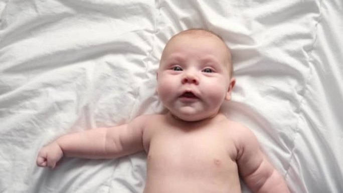 穿着尿布的快乐裸体婴儿躺在白色床单上，抚摸着他的脸。红头发的女婴看着相机。浅色背景。小孩子。严肃的情