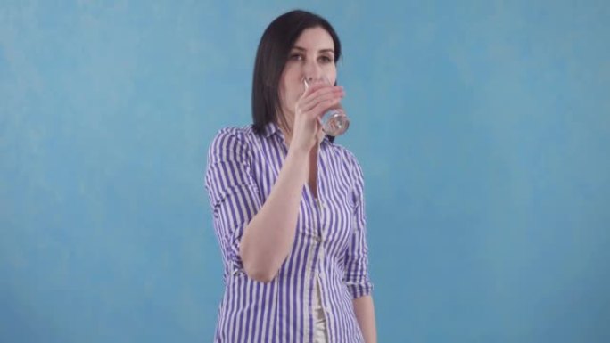 穿着快乐衬衫的年轻女子从站在蓝色背景上的玻璃杯中喝干净的过滤水