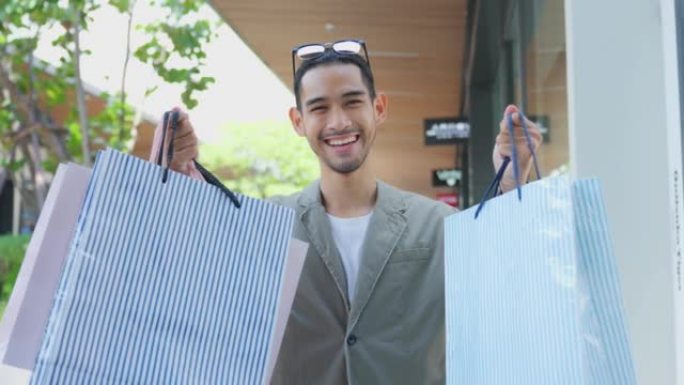百货商店户外亚洲英俊年轻男性购物商品的肖像。迷人的男人拿着购物袋，然后微笑着独自行走，喜欢在购物中心