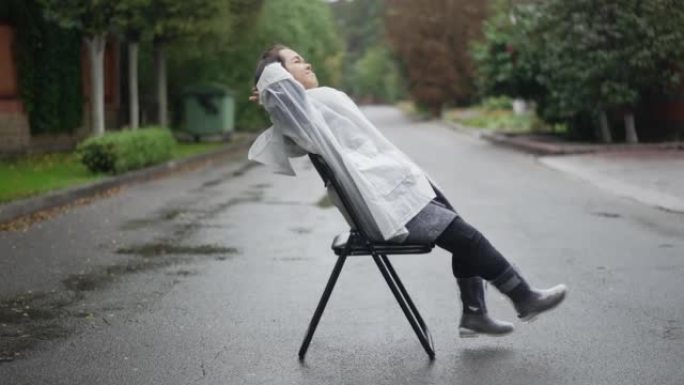 穿着雨衣和橡胶靴的放松小人坐在椅子上微笑着享受郊区阴雨天。自信的高加索小女人在户外路上休息的侧视图。