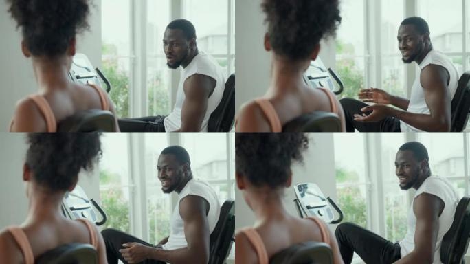 快乐的美国非洲夫妇在开始骑静态自行车的时候一起聊天，面对着健身房外面美丽的镜子窗户。有吸引力的运动人