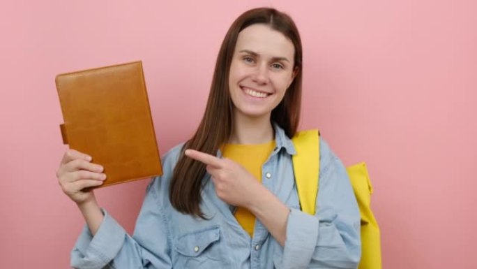 肖像快乐的女孩青少年学生用手指指着书，露出拇指，穿着黄色背包，在粉色背景工作室中孤立地摆姿势。高中教