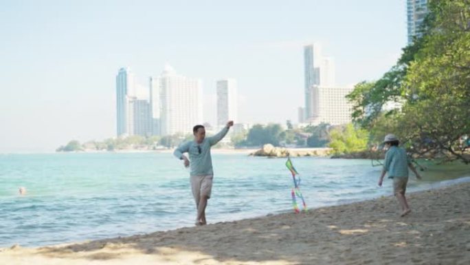 亚洲父亲在沙滩上玩五颜六色的风筝，而他开朗的小儿子则随风飘动着风筝奔跑。他们很开心，有笑脸。假期与家