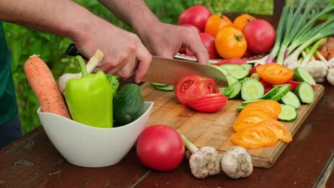 男人在户外烹饪蔬菜沙拉。在花园特写中，男人的手用刀在木板上切红色西红柿。健康饮食，节食，素食，有机夏