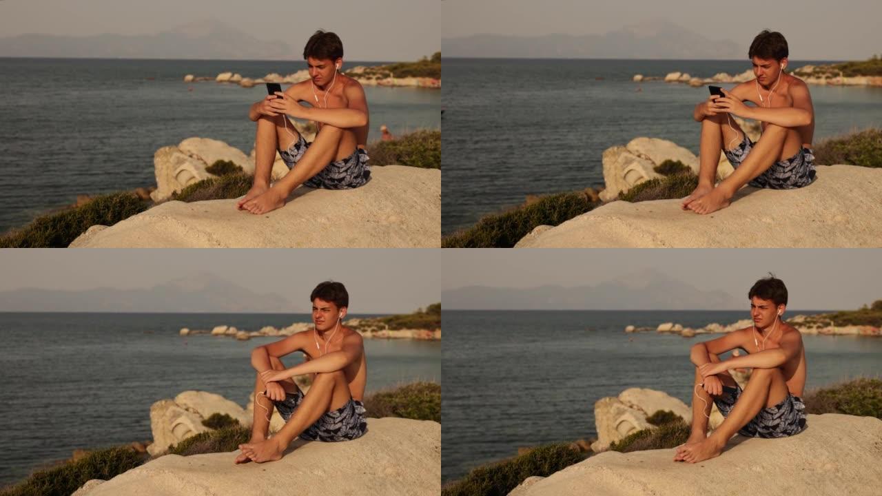 白人十几岁的男孩坐在海边的岩石上听音乐