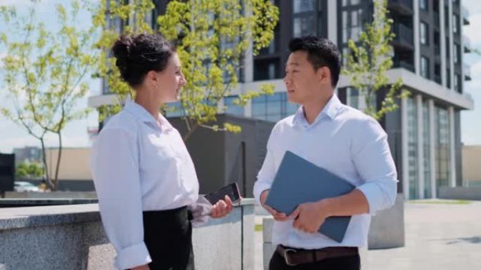 亚洲族裔商人站在办公楼附近，与漂亮的女人讨论合作项目。中国男子拿着笔记本电脑在户外与女同事交谈。