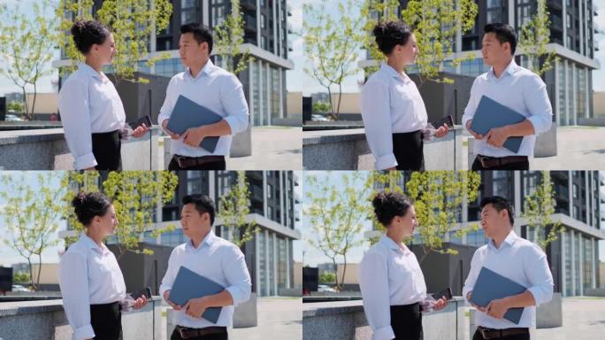 亚洲族裔商人站在办公楼附近，与漂亮的女人讨论合作项目。中国男子拿着笔记本电脑在户外与女同事交谈。