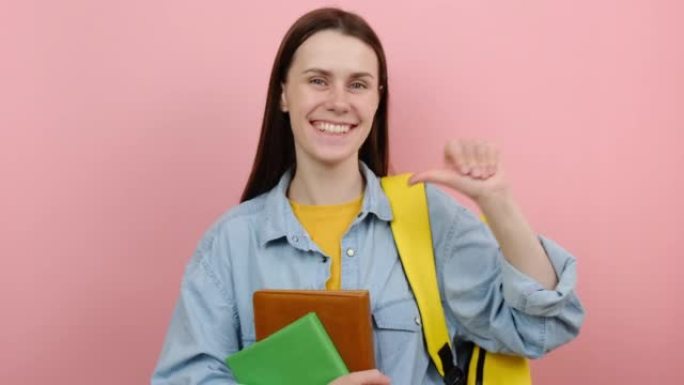 开朗可爱的女孩十几岁的学生穿着衬衫和黄色背包，用手指指着自己的书，在粉色背景工作室中孤立地摆姿势。大