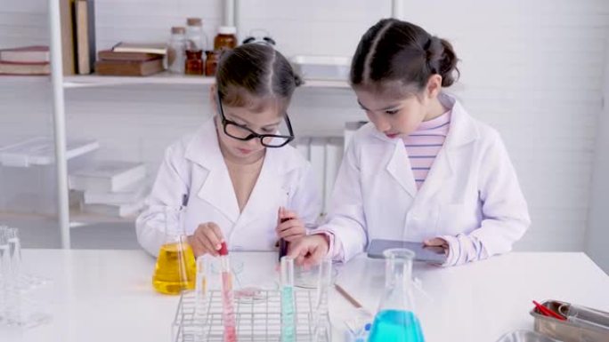 4K，两个亚洲女孩，站在科学教室，混合颜料，做化学实验，用挤压管挤出彩色水滴到测试杯，另一个人拿着笔