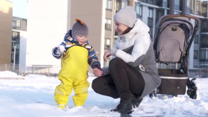 妈妈带着婴儿在冬天穿着温暖的雪衣，冬天在操场上给孩子们穿黄色防水雨围兜，一岁的男婴在寒冷的季节散步。