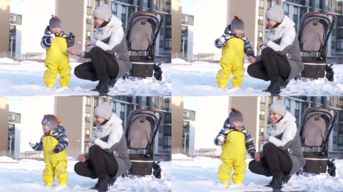 妈妈带着婴儿在冬天穿着温暖的雪衣，冬天在操场上给孩子们穿黄色防水雨围兜，一岁的男婴在寒冷的季节散步。