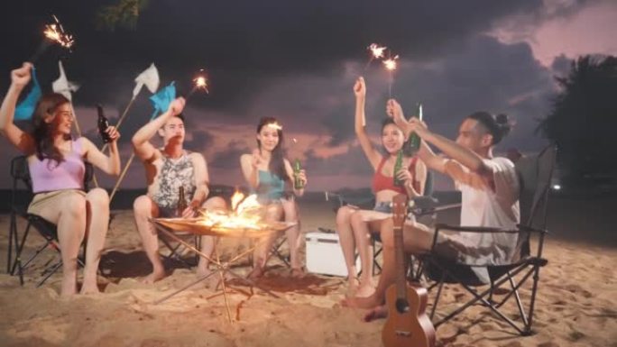 一群亚洲年轻男女晚上在海滩上聚会。迷人的朋友旅行者在海边露营时唱歌跳舞，一起享受热带海岛的假期旅行