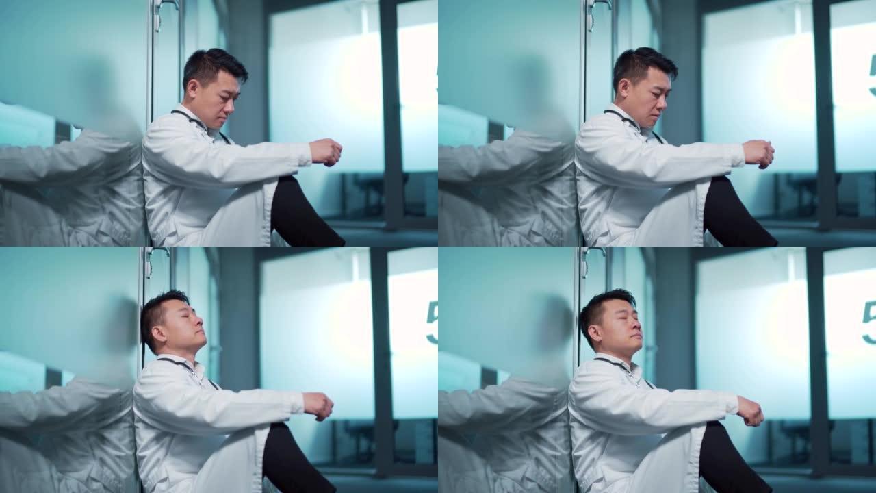 紧张疲惫的亚洲医生坐在医院的走廊里。疲倦沉思的医生在临床上悲伤的男性压力的医护人员