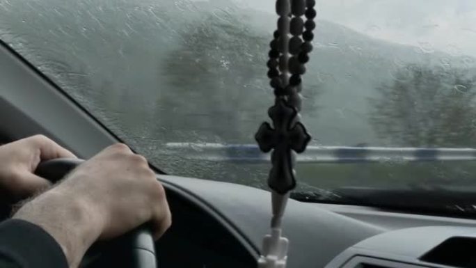 大雨中，一名男子用强壮的双手握住方向盘，用雨刷清洁挡风玻璃上的水。车里挂着一个亚美尼亚十字架形状的护