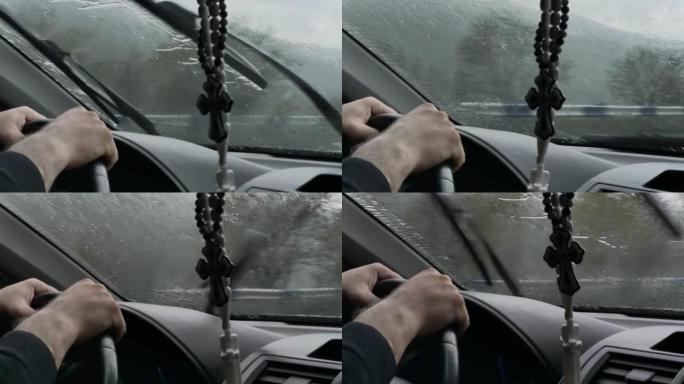 大雨中，一名男子用强壮的双手握住方向盘，用雨刷清洁挡风玻璃上的水。车里挂着一个亚美尼亚十字架形状的护