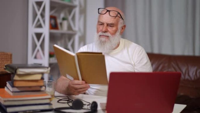 集中的高年级男子在笔记本电脑上微笑着在线阅读图书消息。留着长胡子和白发的白人男性退休人员的肖像在室内